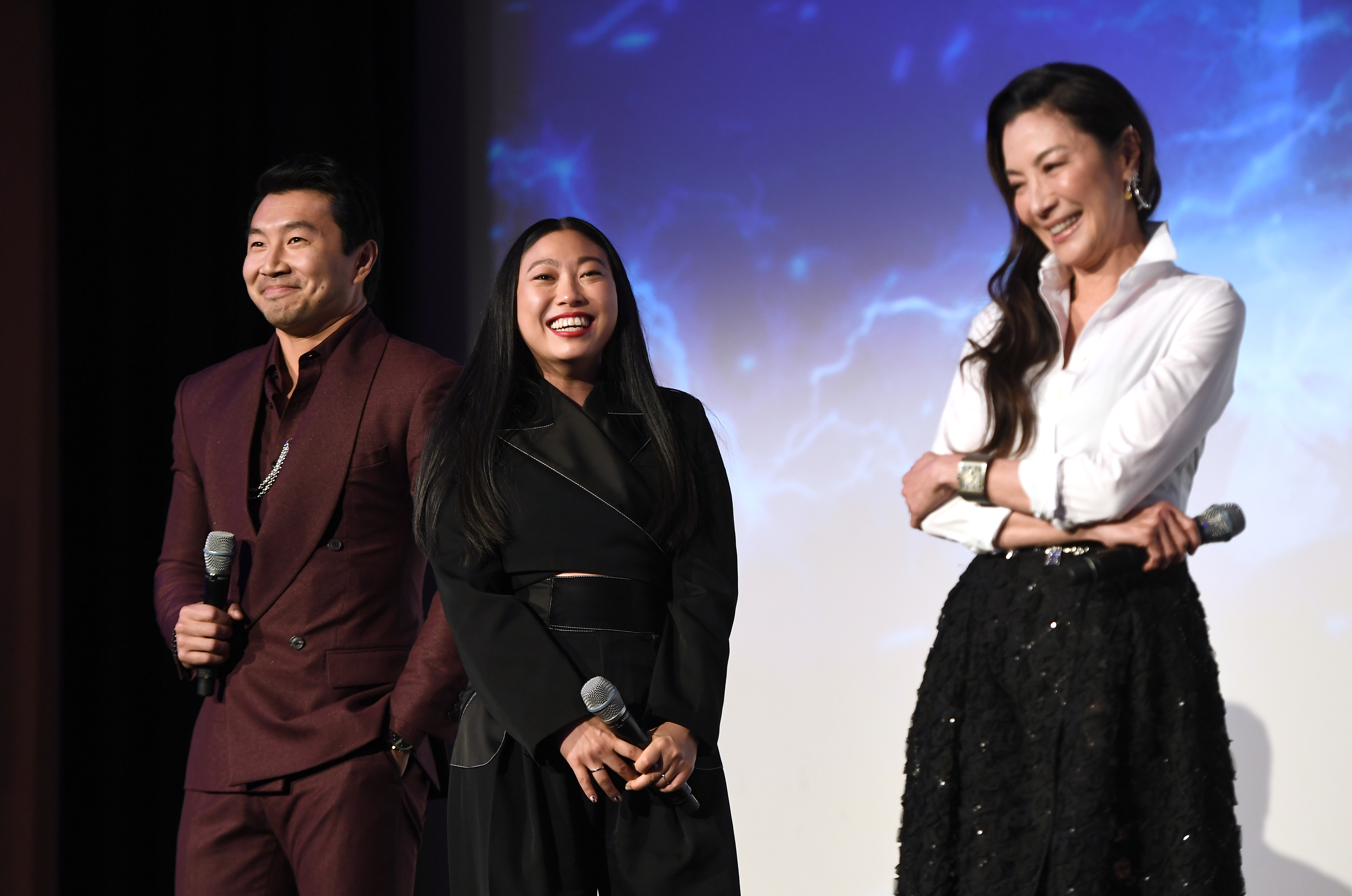 'Shang-Chi' stars Simu Liu, Awkwafina, and Michelle Yeoh hold microphones and speak onstage before the movie hits the box office. Liu wears a maroon suit, Awkwafina wears a black jacket with a black belt, and Yeoh wears a black sparkly skirt and a white button down shirt.