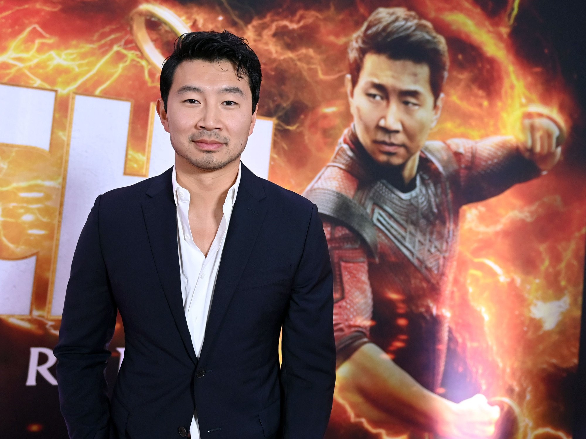 'Shang-Chi and the Legend of the Ten Rings' star Simu Liu. He's wearing a white shirt and black jacket and is standing in front of a picture of his character, Shang-Chi. Could his Marvel movie get a spinoff?