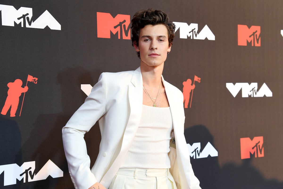 Shawn Mendes wearing a white suit jacket over a white tank top and pants, posing at an event.