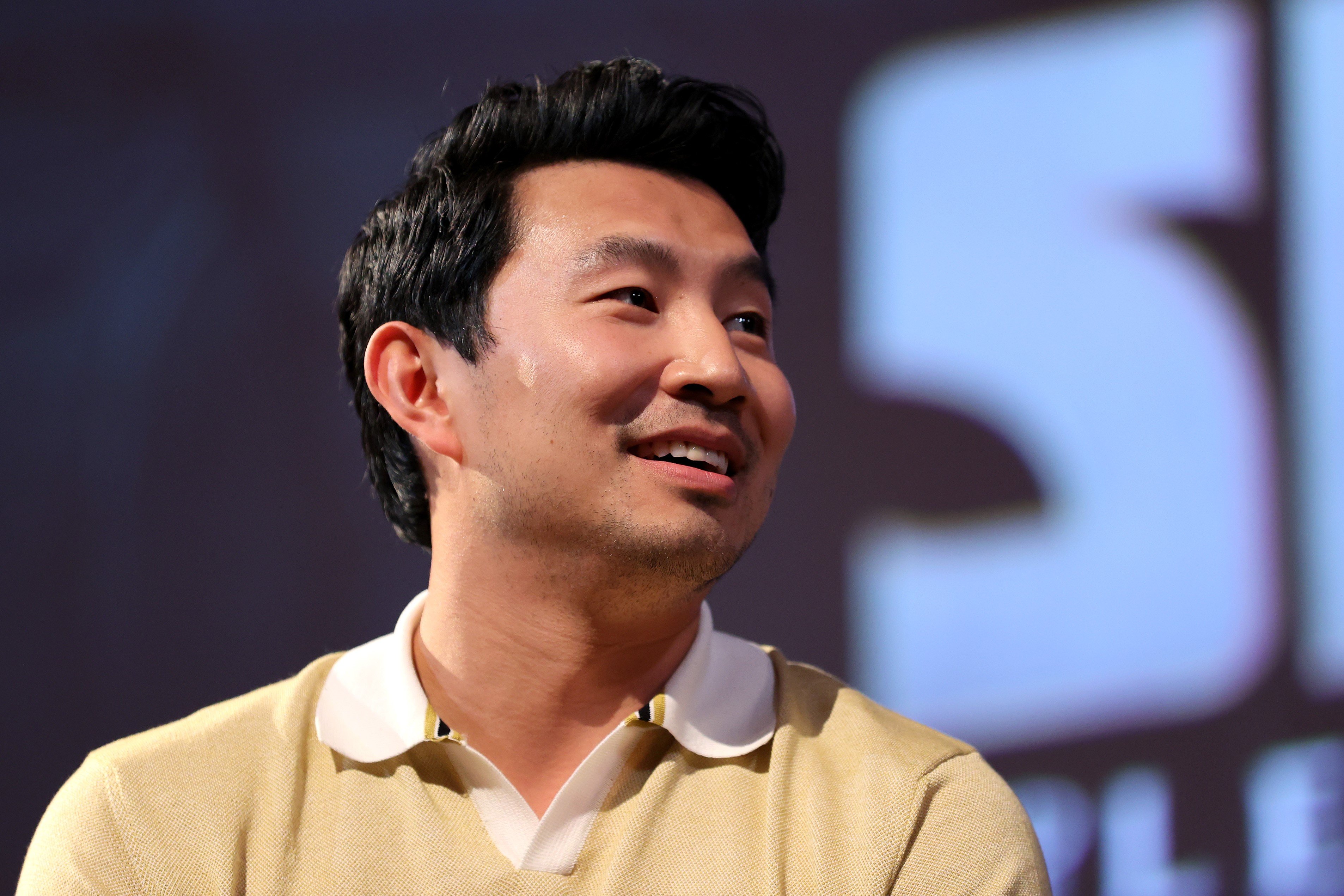 Simu Liu speaking during an event for Marvel's Shang-Chi and the Legend of the Ten Rings. He is wearing a yellow shirt and is looking toward his left.