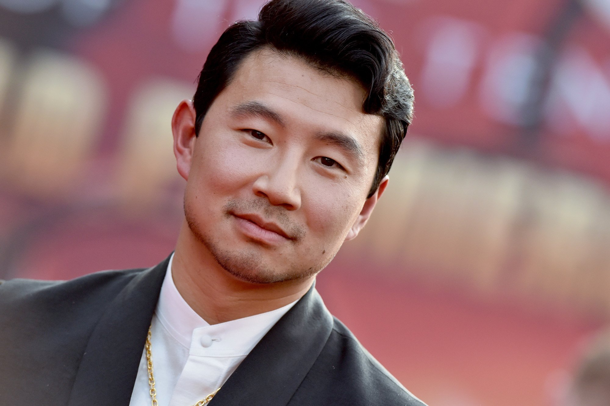 'Shang-Chi' star Simu Liu. He's wearing a white shirt, black jacket, and golden chain. He's smiling at the camera, and a blurred red wall is behind him. This photo is for our article questioning, when is 'Shang-Chi' coming to Disney+?