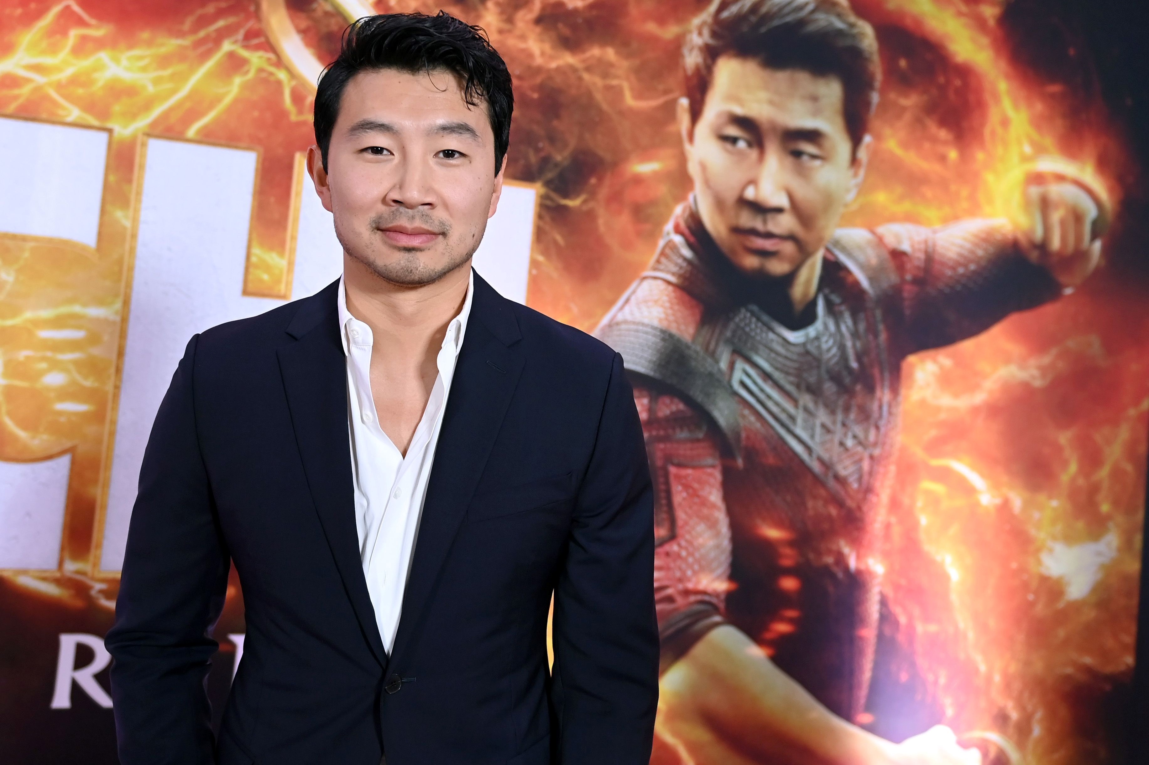 Actor Simu Liu standing in a white shirt and black jacket from waist up in front of his poster from the Marvel movie 'Shang-Chi and the Legend of the Ten Rings.'