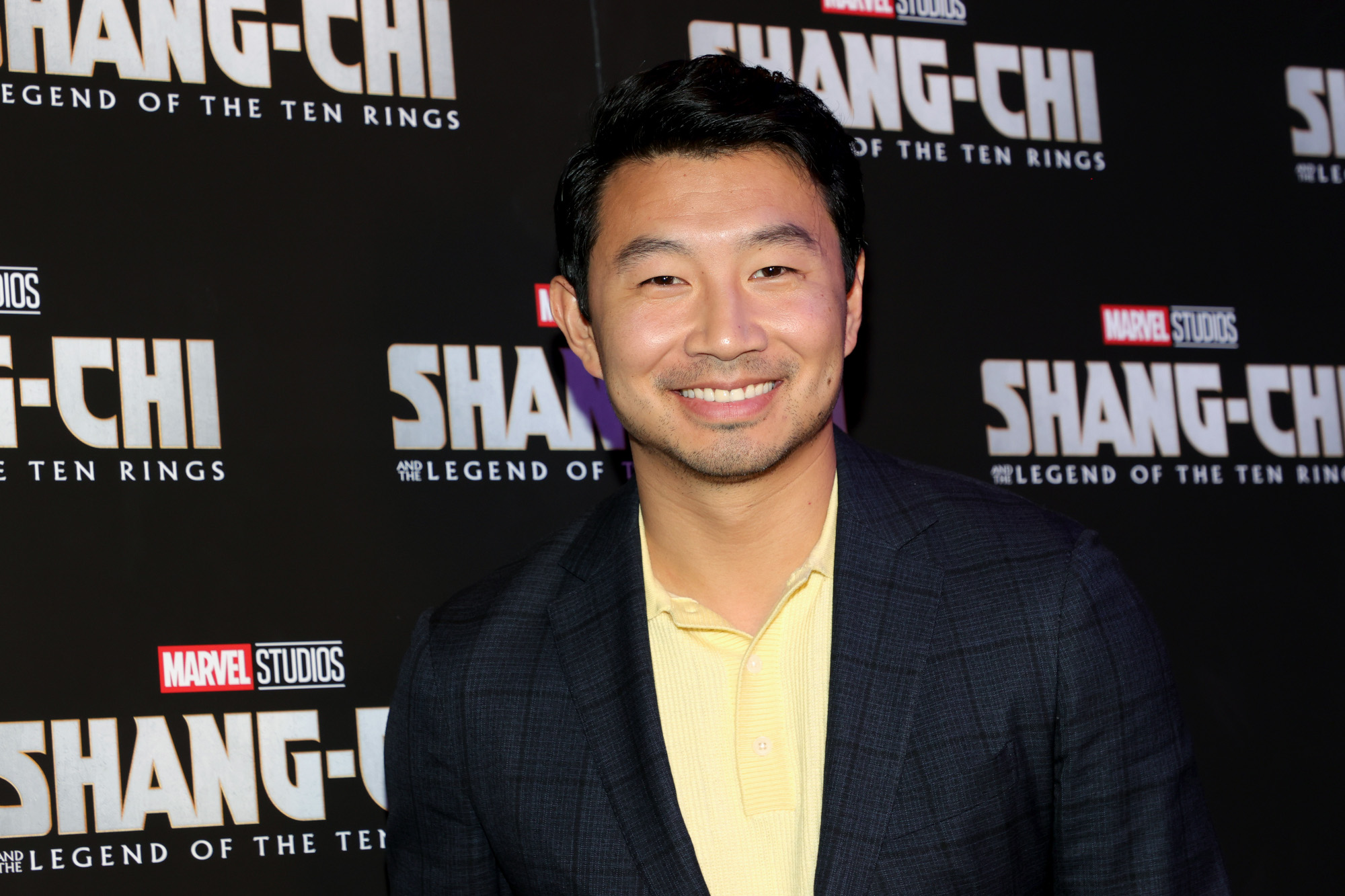 'Shang-Chi and the Legend of the Ten Rings' star Simu Liu. He's wearing a yellow button-up shirt and black jacket. He's smiling at the camera and standing in front of a wall with the 'Shang-Chi' logo all over it.