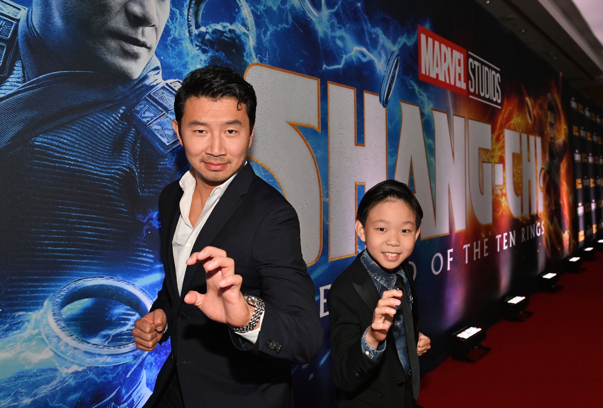 'Shang-Chi and the Legend of the Ten Rings' stars Simu Liu and Jayden Zhang posing in front of a Shang-Chi wall. They're both wearing black suits and have one arm extended making a claw motion.