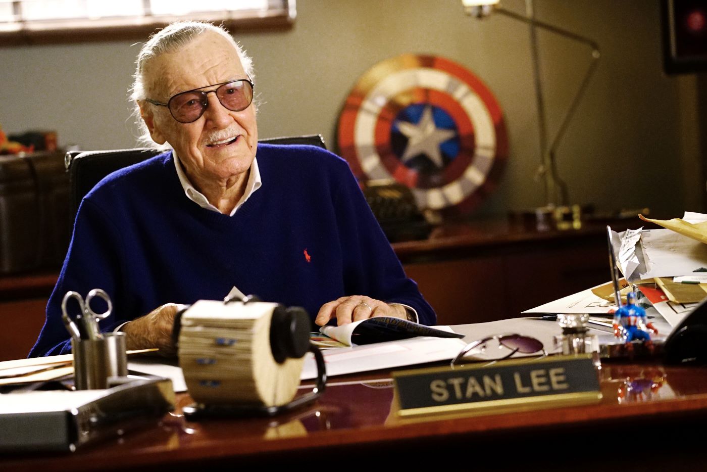 Stan Lee sitting at his desk with marvel memorabilia around him.
