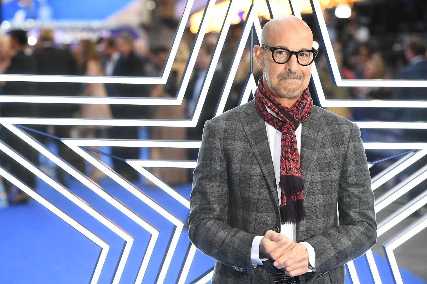 Stanley Tucci dressed in a black and grey plaid suit standing in front of a metal star with multiple layers of the shape from big to small.