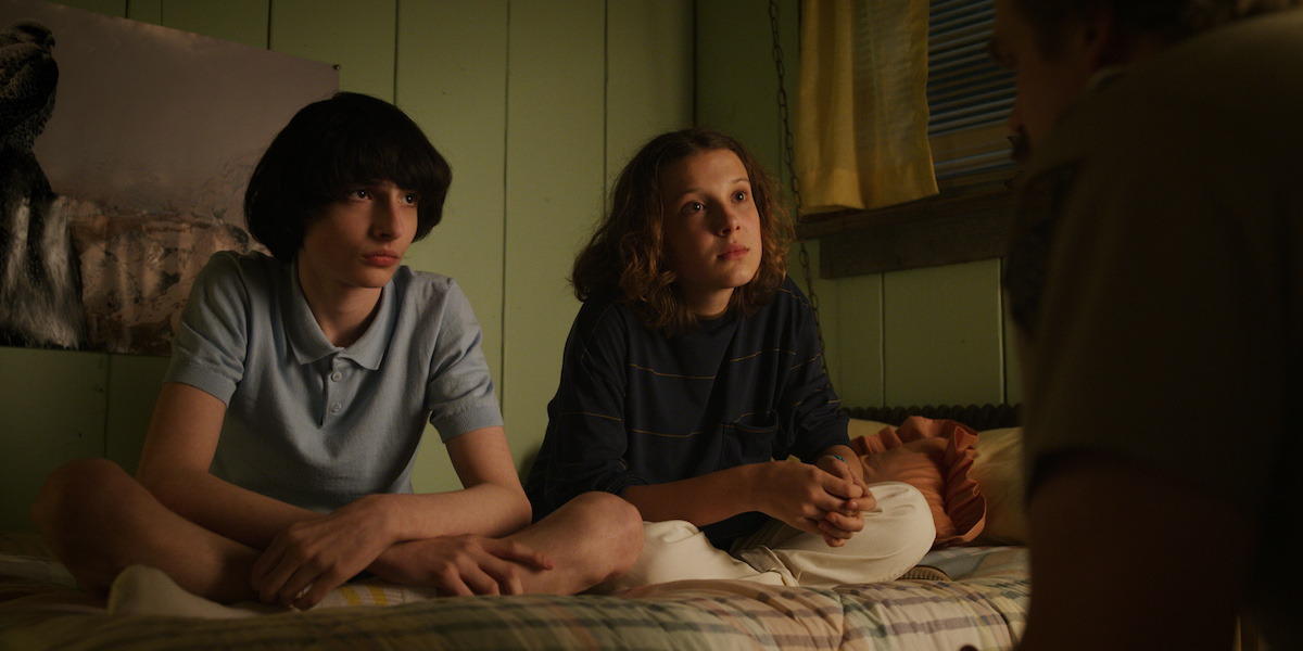 Mike Wheeler, played by Finn Wolfhard, sitting on a bed with Eleven, played by Millie Bobby Brown, in a production still from 'Stranger Things' Season 3.