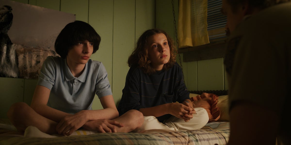 Mike Wheeler, played by Finn Wolfhard, sitting on a bed with Eleven, played by Millie Bobby Brown, in a production still from 'Stranger Things' Season 3.