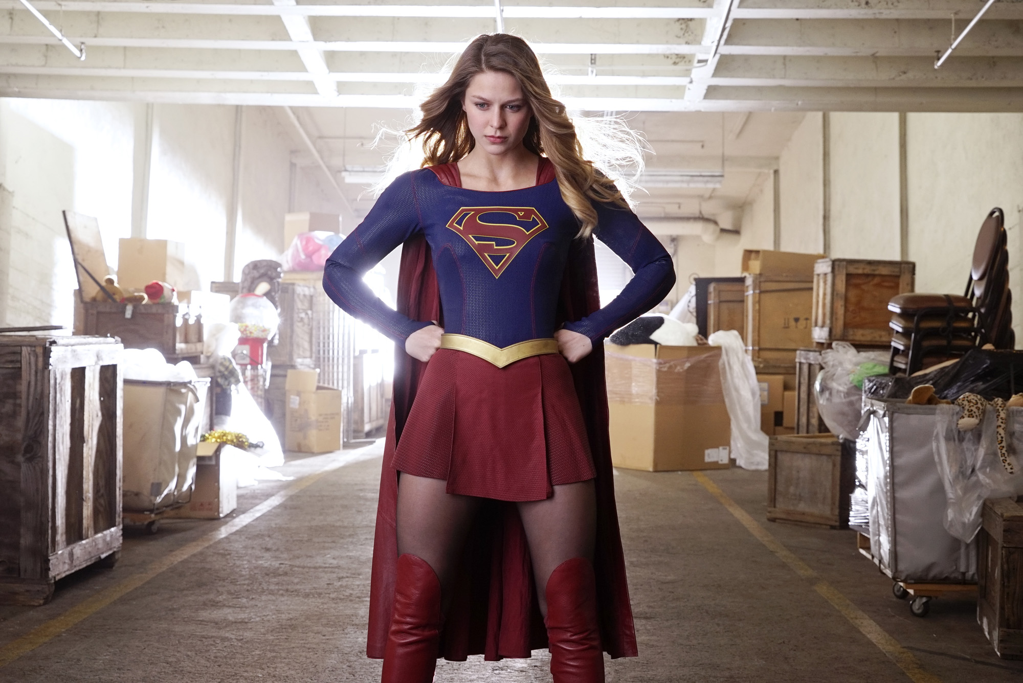 'Supergirl' Season 6 Episode 13 star Melissa Benoist poses in her Supergirl costume with her hands on her hips. The suit has a red skirt, red boots, red cape, and a blue long-sleeved shirt with the House of El crest on it.