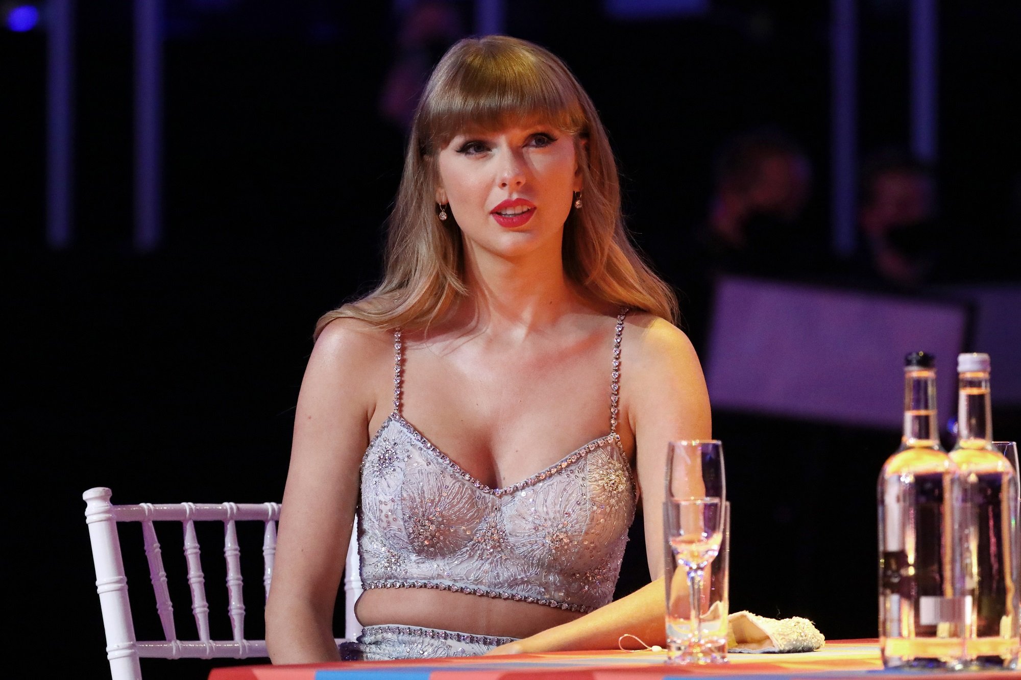 Taylor Swift wears a beaded top while sitting at a table