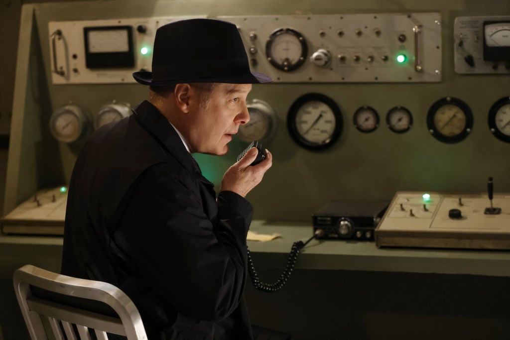 James Spader as Raymond 'Red' Reddington speaks into a transponder from the Latvia sit. He's wearing a dark suit and fedora.