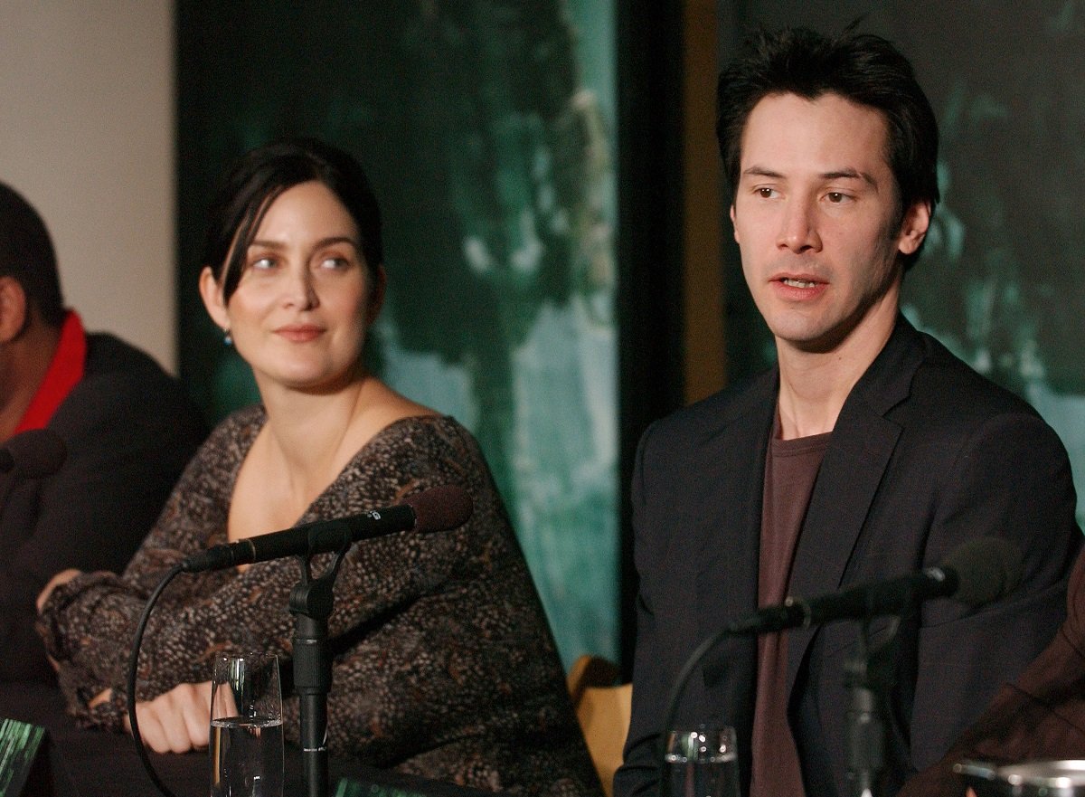 Carrie-Anne Moss looks at Keanu Reeves as he speaks into a microphone.