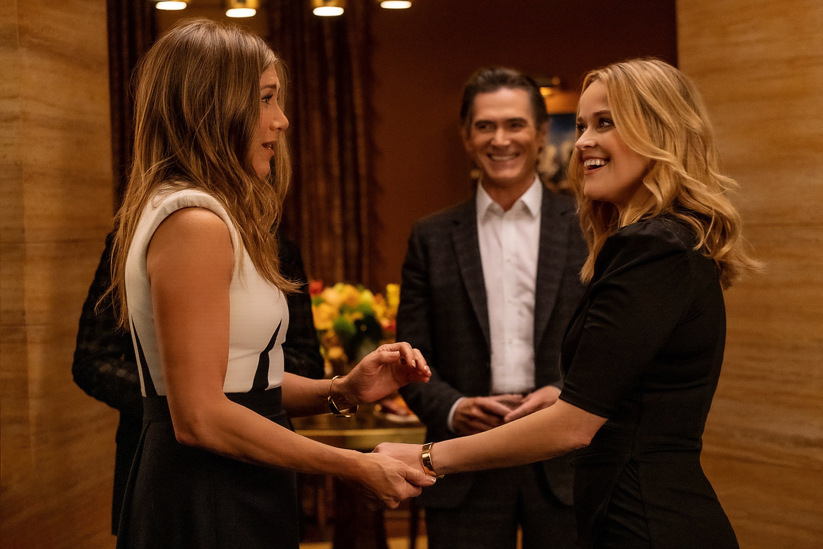 The Morning Show stars Jennifer Aniston and Reese Witherspoon hold hands as Billy Crudup smiles in a scene from season 2