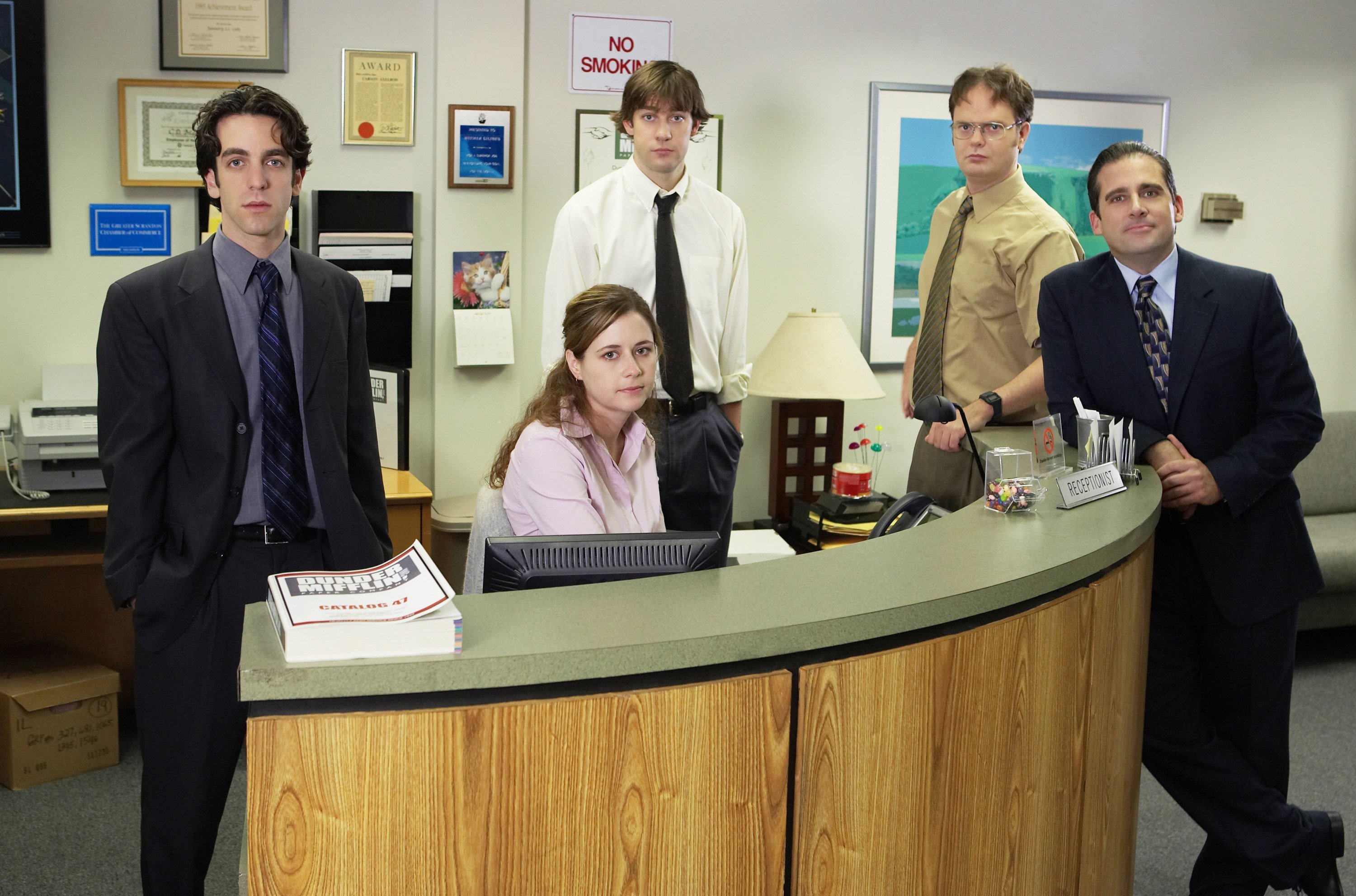 'The Office' cast posing at the reception desk, including BJ Novak as Ryan Howard, Jenna Fischer as Pam Beesly, John Krasinski as Jim Halpert, Rainn Wilson as Dwight Schrute, and Steve Carell as Michael Scott