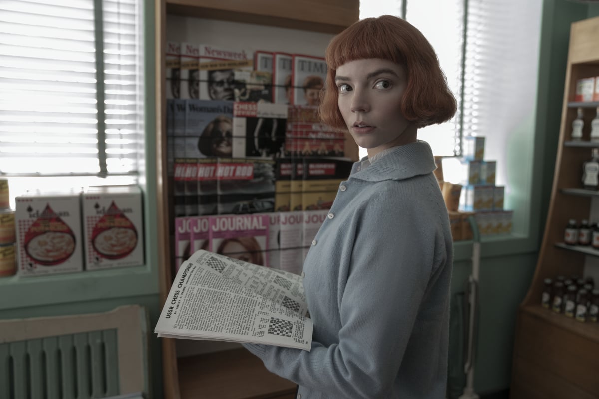 Beth Harmon holds a newspaper and looks to the side. She has blunt bangs and is wearing a light blue sweater.