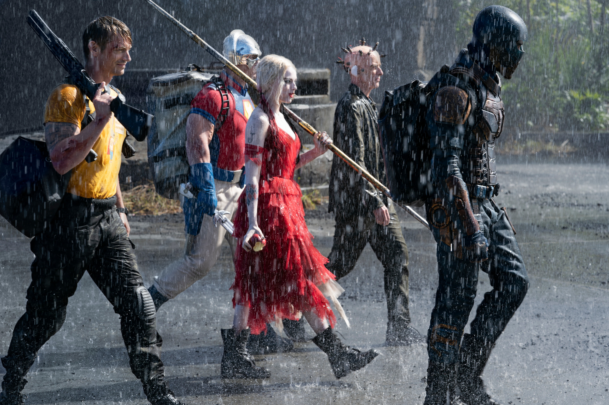 Joel Kinnaman as Rick Flag, John Cena as Peacemaker, Margot Robbie as Harley Quinn, Peter Capaldi as The Thinker and Idris Elba as Bloodsport in ‘The Suicide Squad.’ The group walks together in the rain, headed somewhere to the right of screen. Bloodsport leads them wearing all black and a black helmet, and Harley Quinn wears a ruffled red dress and combat boots. The film is streaming on HBO Max and available in theaters.