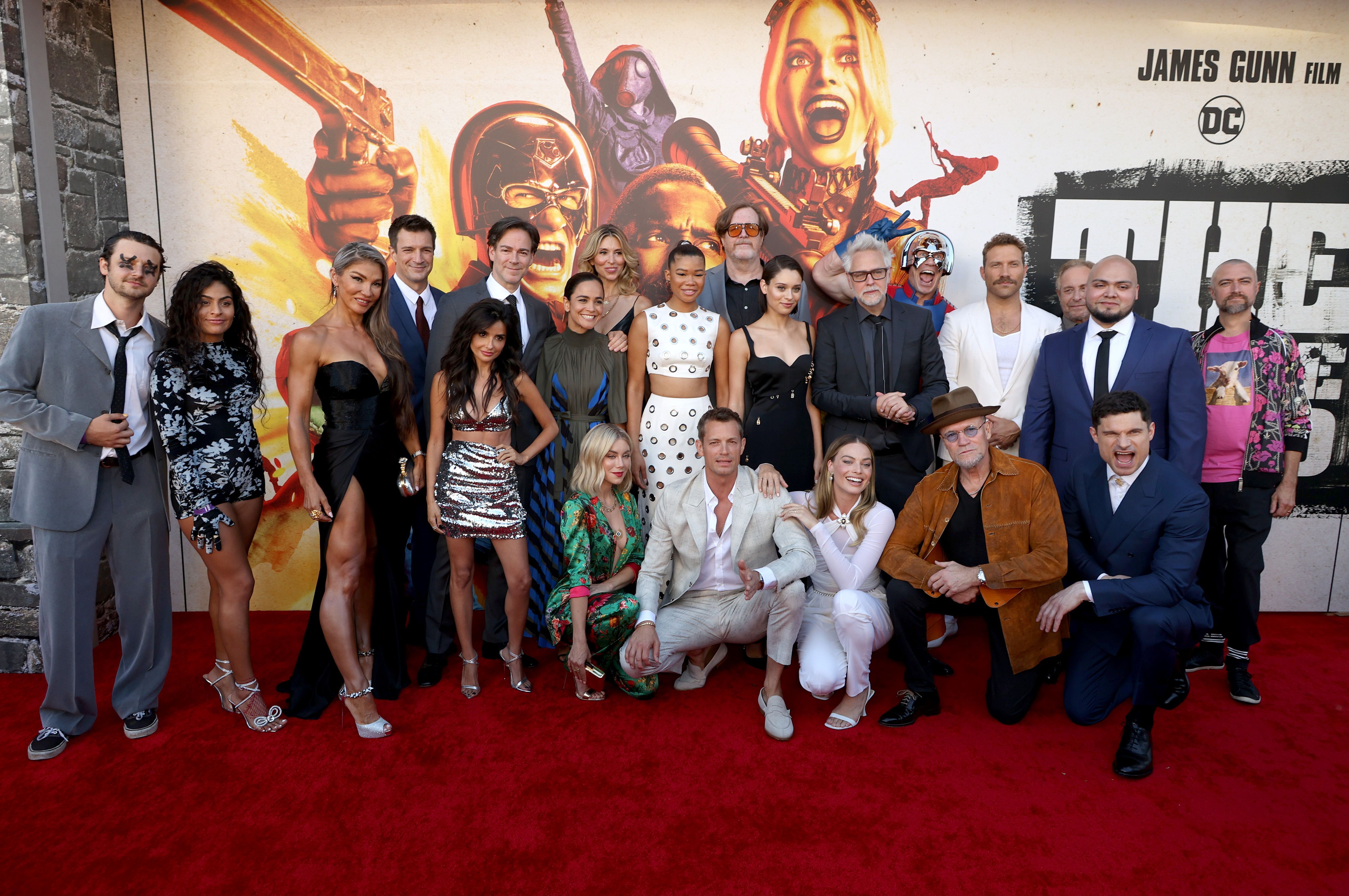 The stars of 'The Suicide Squad' gag reel huddle together to pose in front of a poster for the movie on the red carpet.