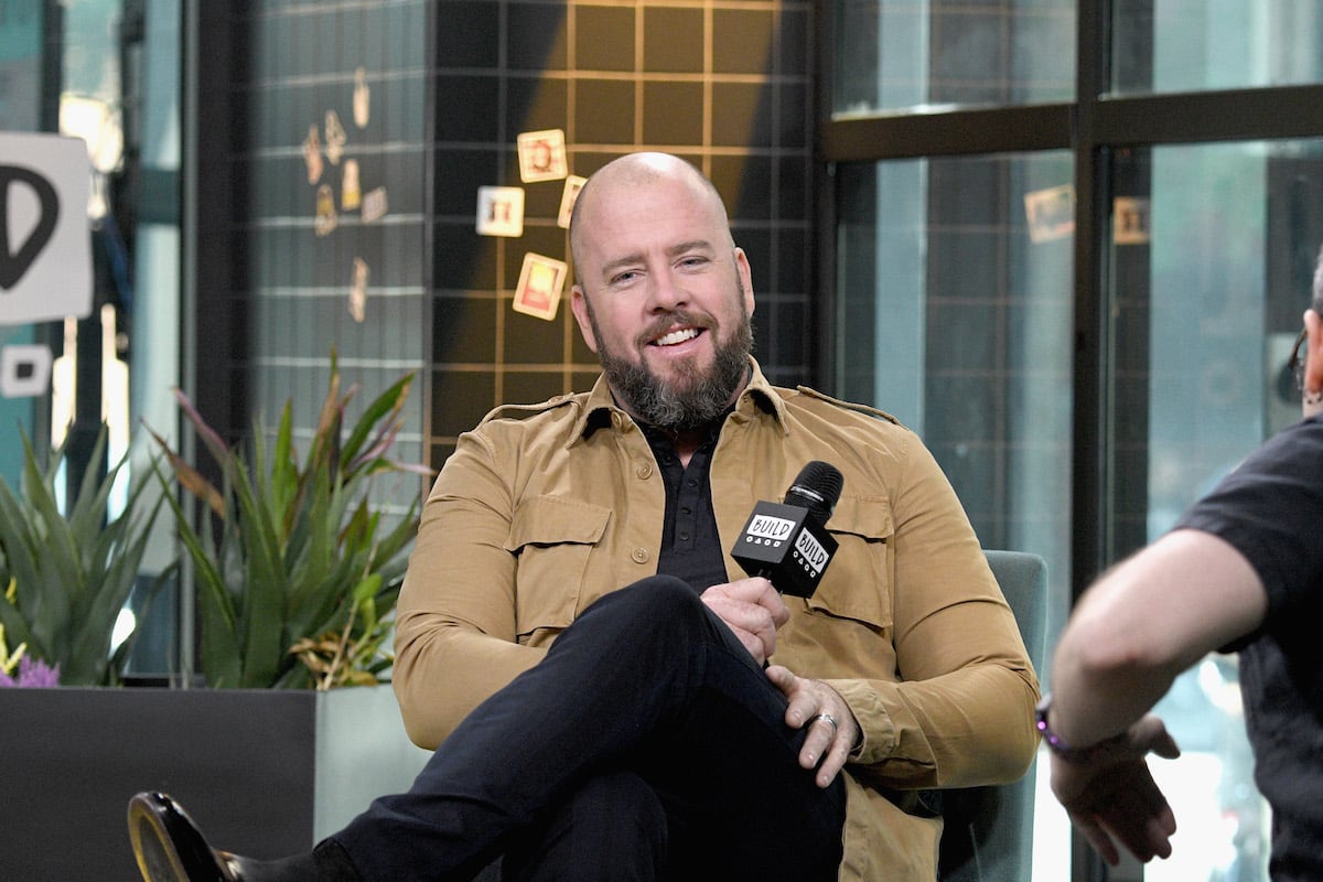 'This Is Us' star Chris Sullivan smiles wearing a brown jacket and holding a microphone during a visit to Build Series in 2019