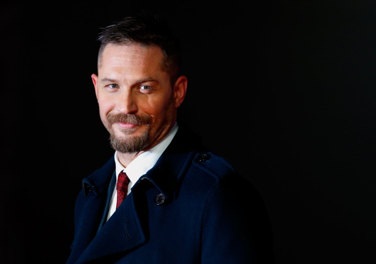 Tom Hardy with a beard and mustache smiling and wearing a blue suit and red tie. 