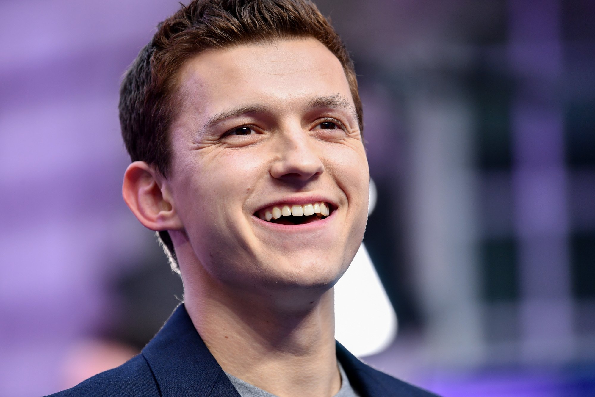 Spider-Man star Tom Holland, who does not appear in episode 5 of Marvel's 'What If...?' The photo shows him from the neck up. He's wearing a dark blue suit and smiling up at something.