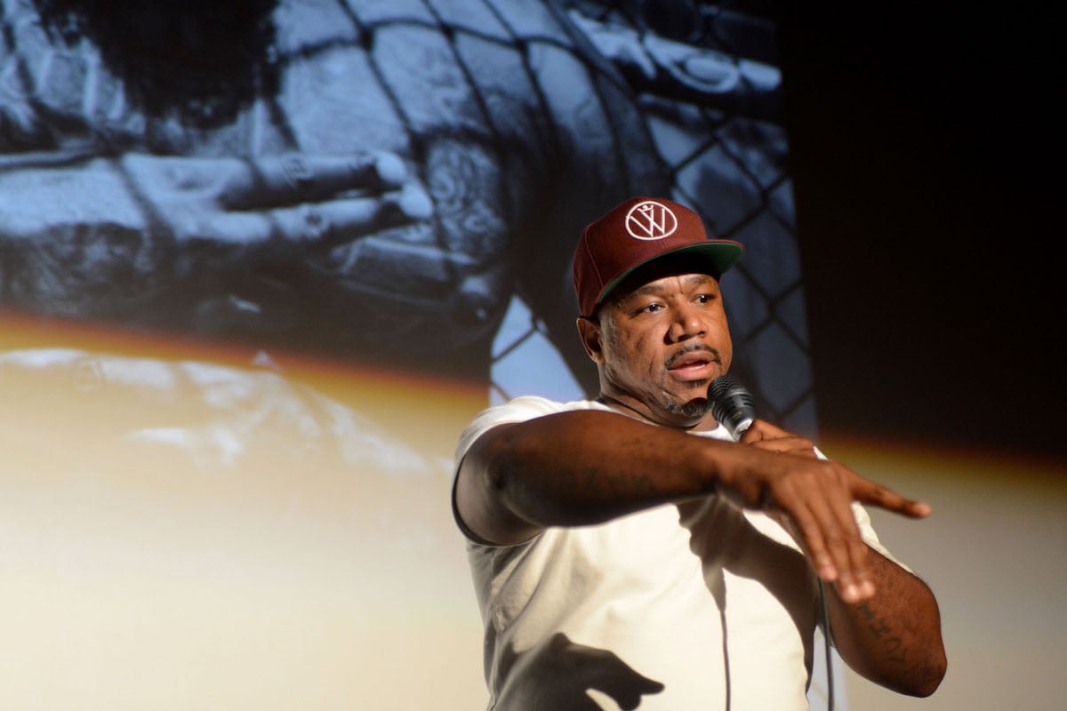Wack 100 wearing a white shirt and maroon hat on stage holding a microphone.
