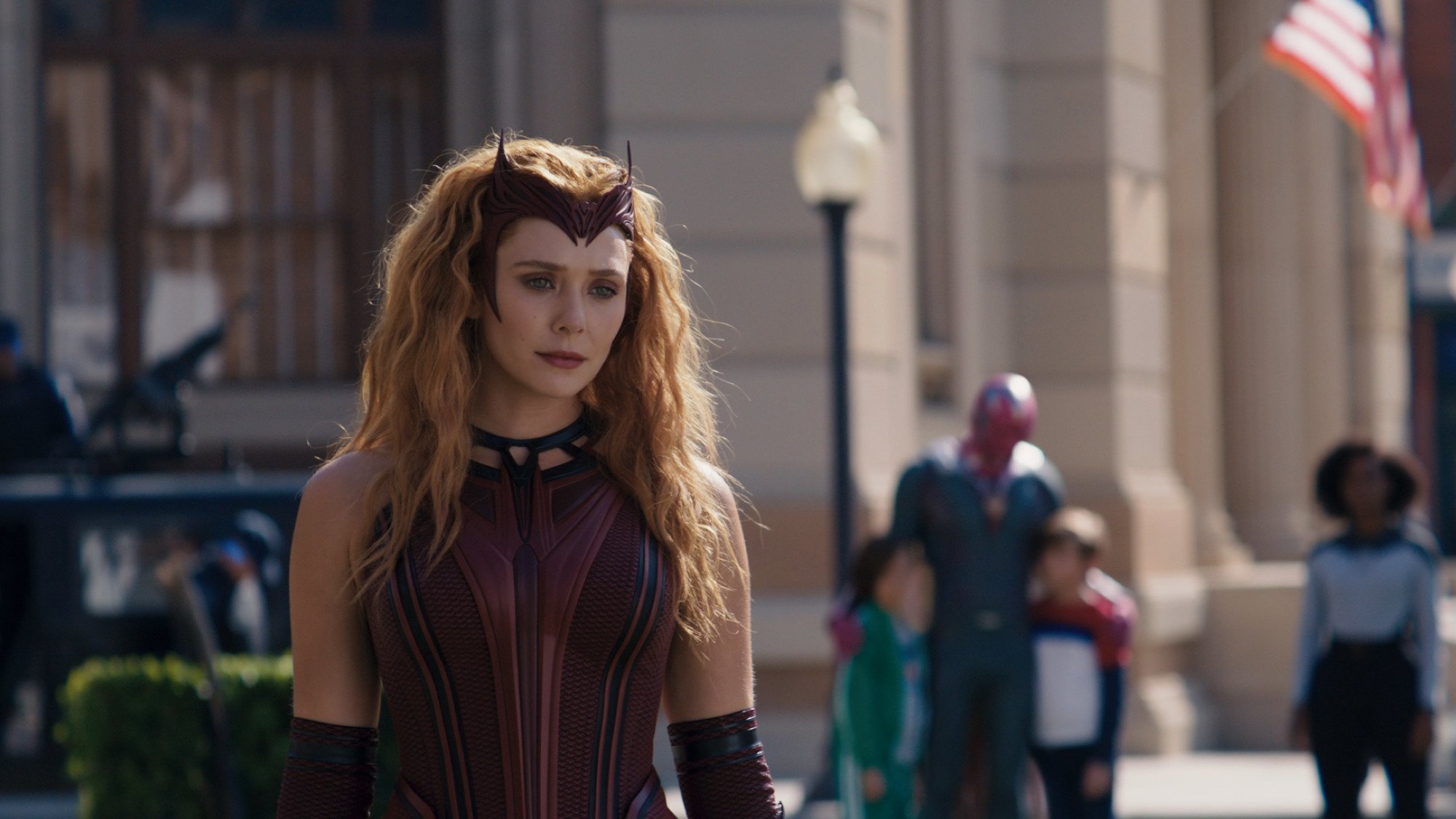 Elizabeth Olsen as Wanda Maximoff wearingher Scarlet Witch costume in 'WandaVision.' She has a dark red corset, dark red gloves that go up to her elbow, and a headband that looks like devil horns. Her red hair is down and curled, and she's looking down at something.