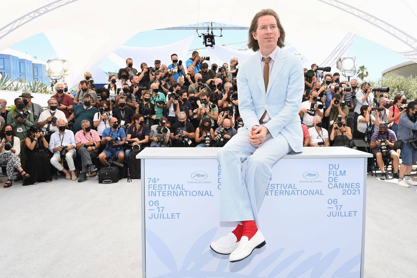 Wes Anderson, director of 'The French Dispatch' sitting on a white box with blue writing dressed in a blue pin stripe suit in front of a masked audience.