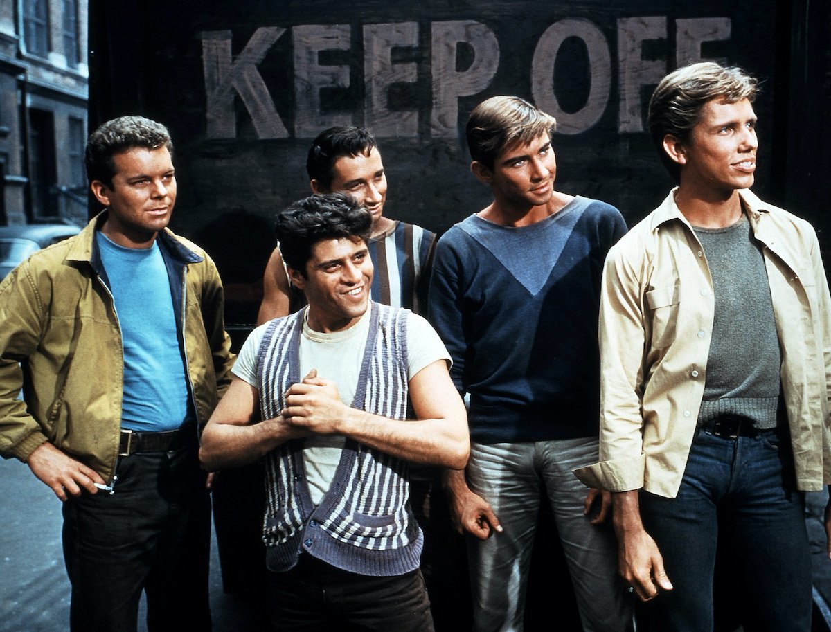 Russ Tamblyn and other members of the 1961 'West Side Story' movie cast stand on a sidewalk with a black wall behind them that says 'KEEP OFF' in bold lettering.