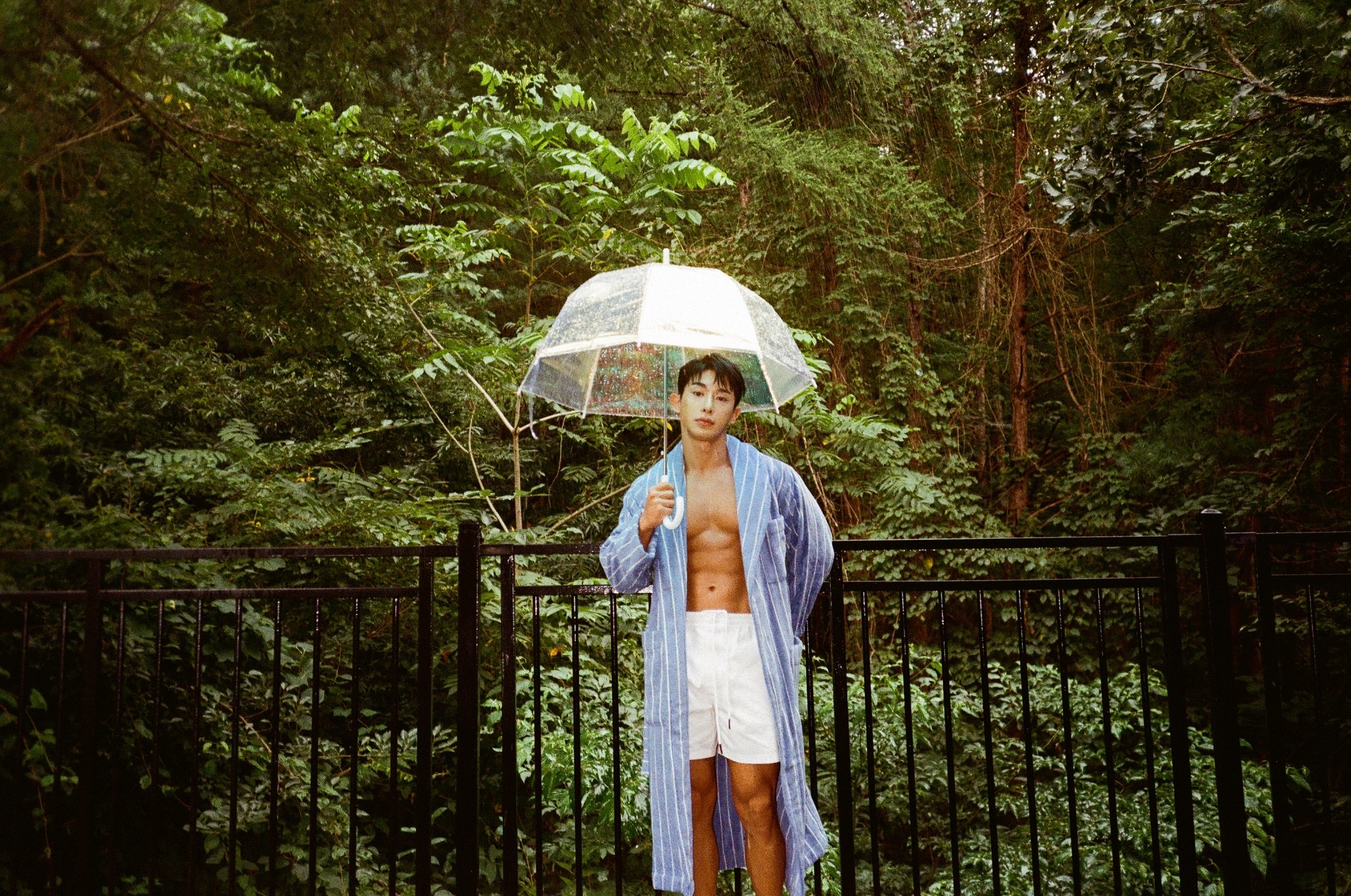 Wonho holds a clear umbrella while wearing a blue-striped robe and white shorts