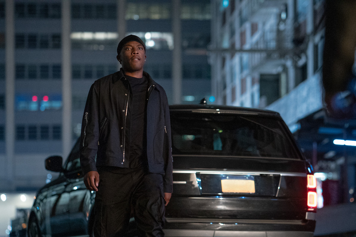 Woody McClain as Cane Tejada dressed in all black standing in front of a car in 'Power Book II: Ghost'