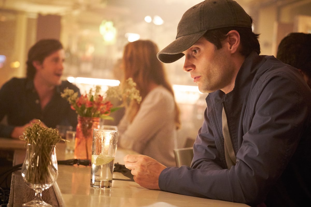 Joe Goldberg sitting at a bar wearing a hat for a disguise.