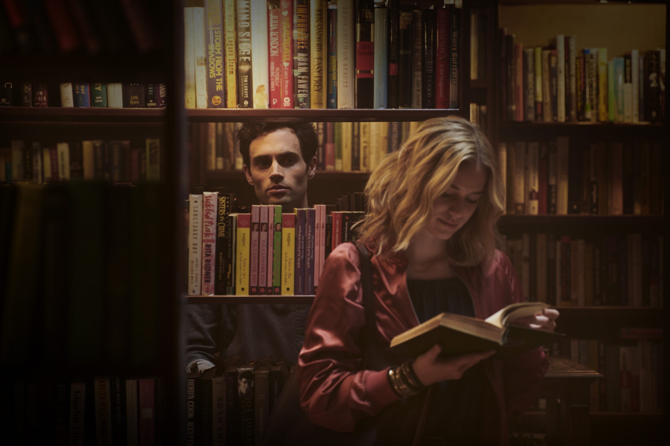 Joe Goldberg stares through a bookshelf at Guinevere Beck reading a book.