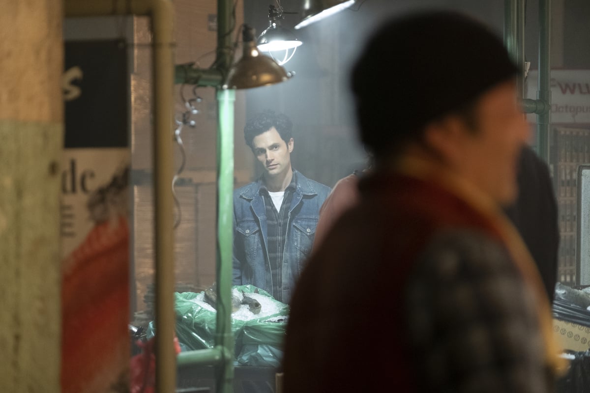 Penn Badgley as Joe Goldberg in 'You' Season 2. Joe watches from afar wearing a plaid shirt and jean jacket.