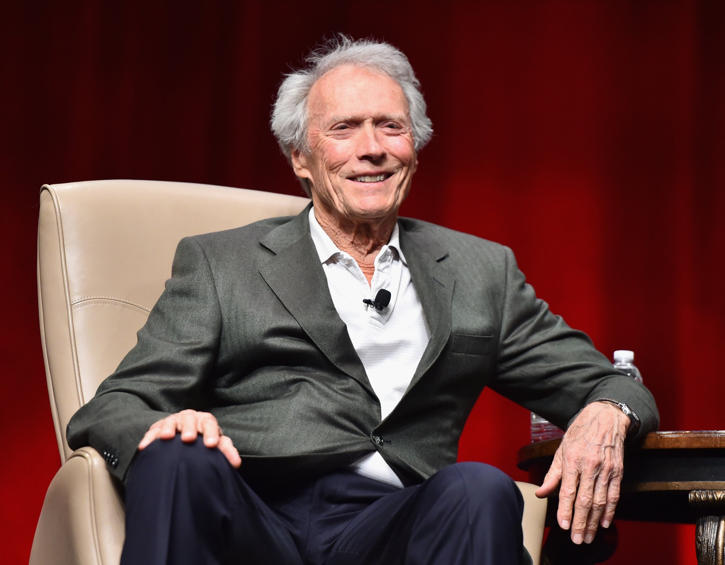 Actor Clint Eastwood sits in a tan leather chair on a stage in front of a red curtain. He wears a grey suit and smiles. The actor has been working in Hollywood for 50 years, and now six Clint Eastwood movies are being re-released in movie theaters to celebrate.