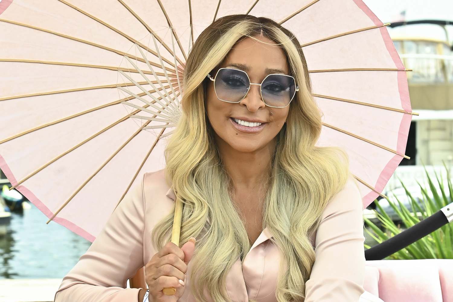 Karen Huger smiles, wears shades with a blue tint, and holds a paper umbrella
