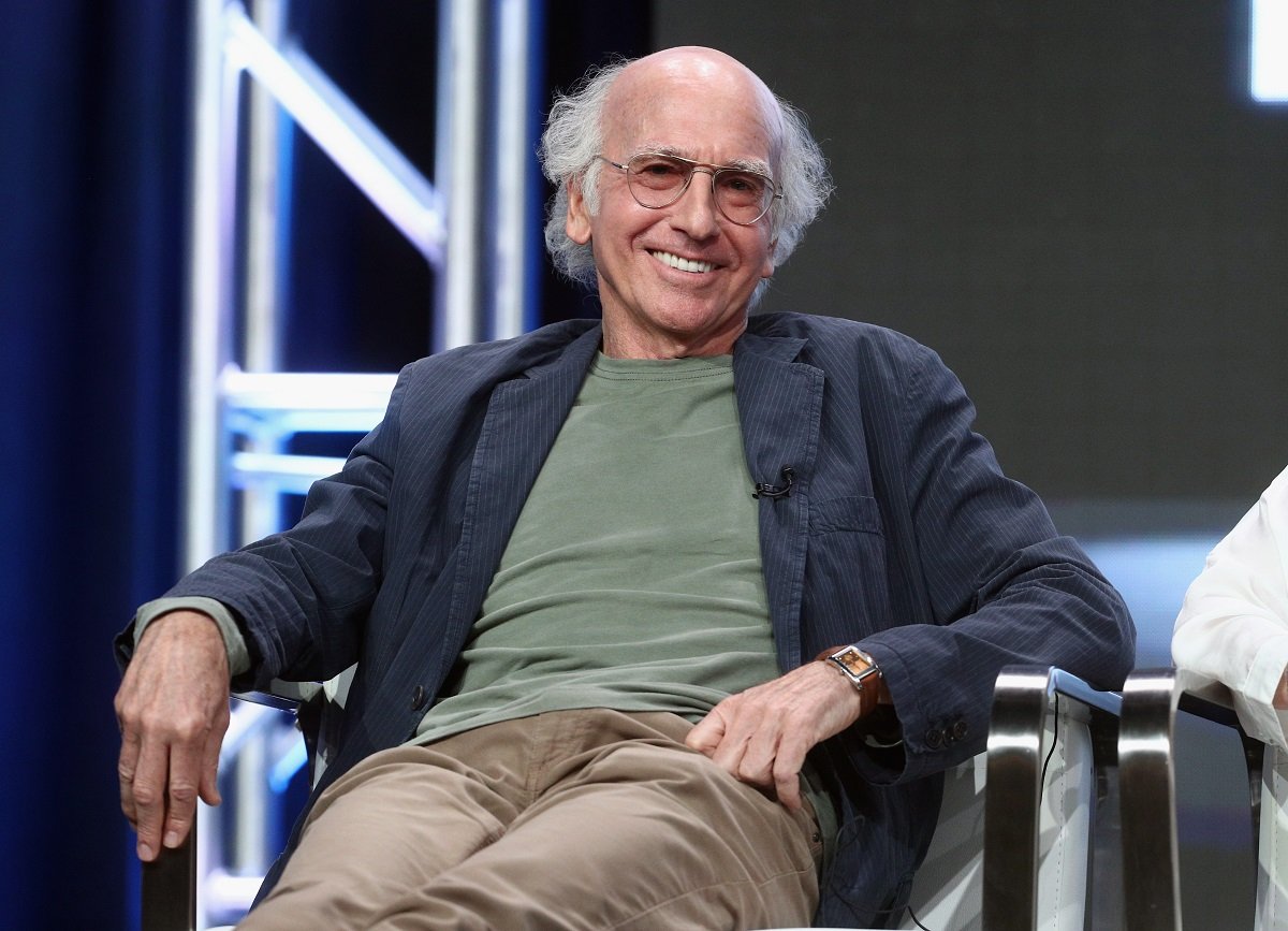 Larry David of 'Curb Your Enthusiam' Season 11, reclining in a chair wearing khakis, a green shirt, and a blue overcoat.