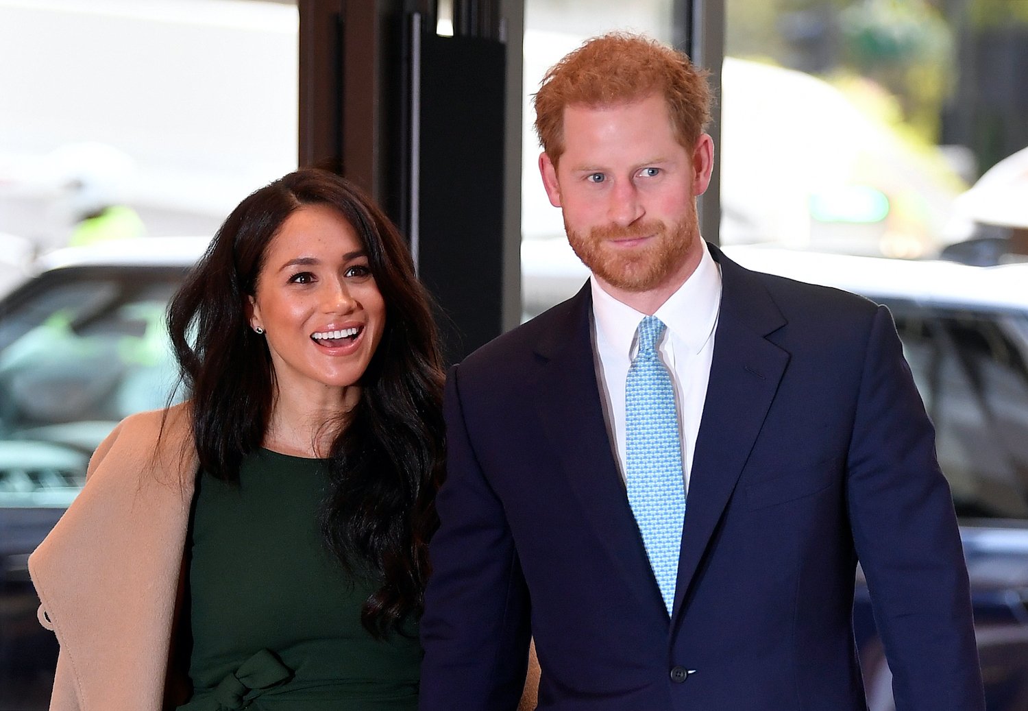 Meghan Markle and Prince Harry smile as they walk together, with Meghan wearing a green dress and brown coat and Prince Harry wearing a blue suit and tie at WellChild Awards