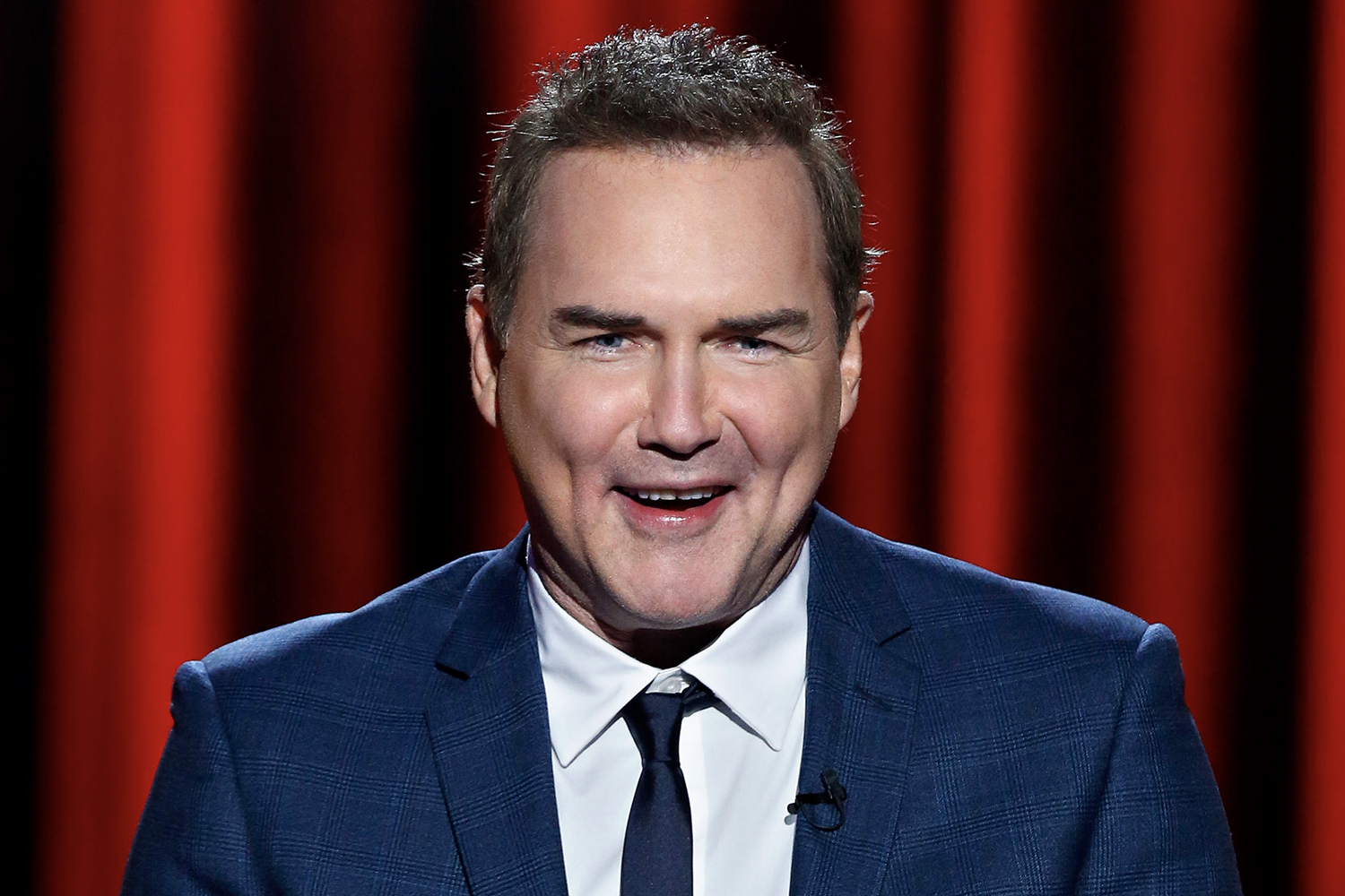 Norm Macdonald smiles standing in front of a red curtain backdrop