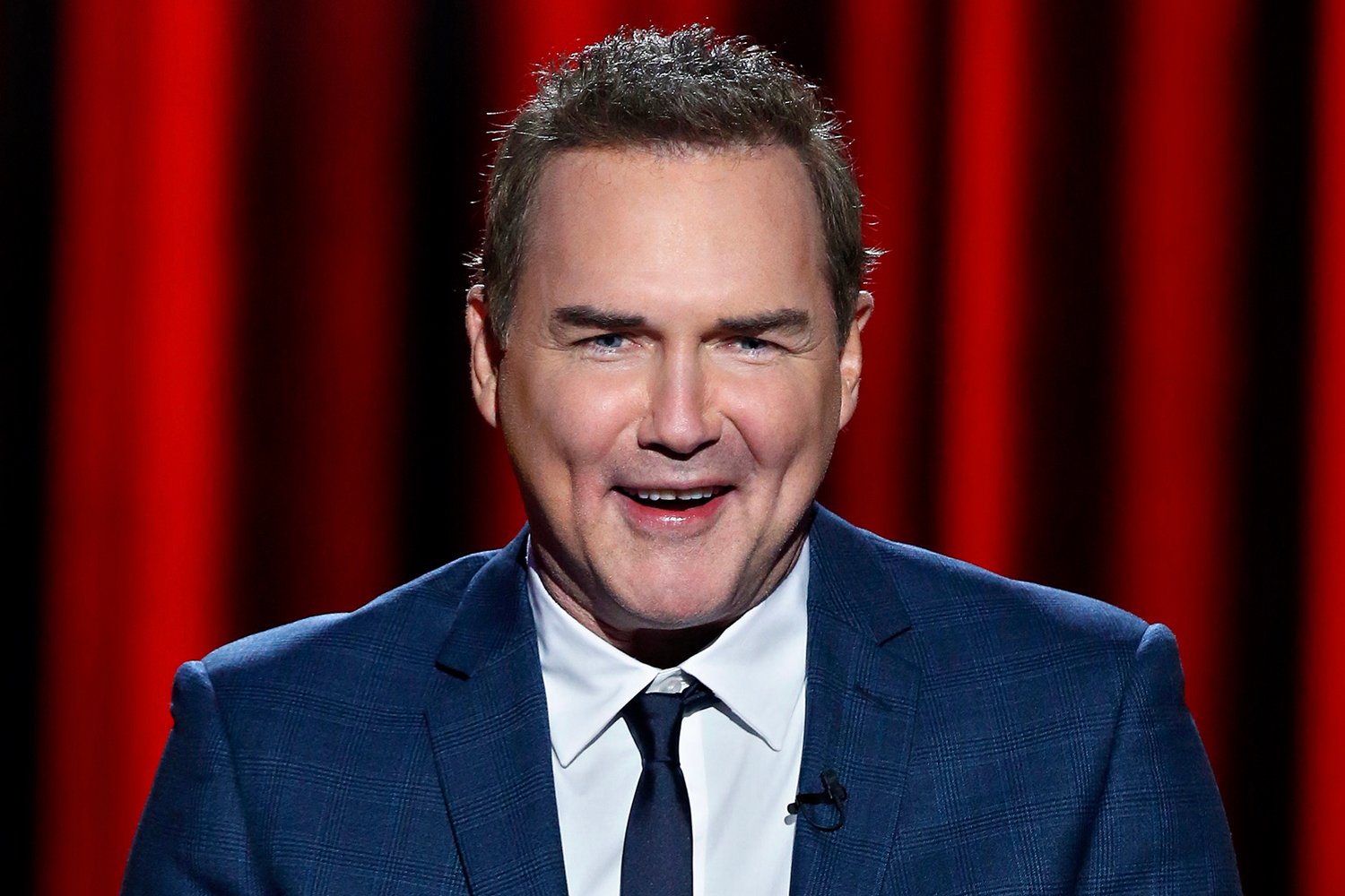 Norm Macdonald smiles standing in front of a red curtain backdrop