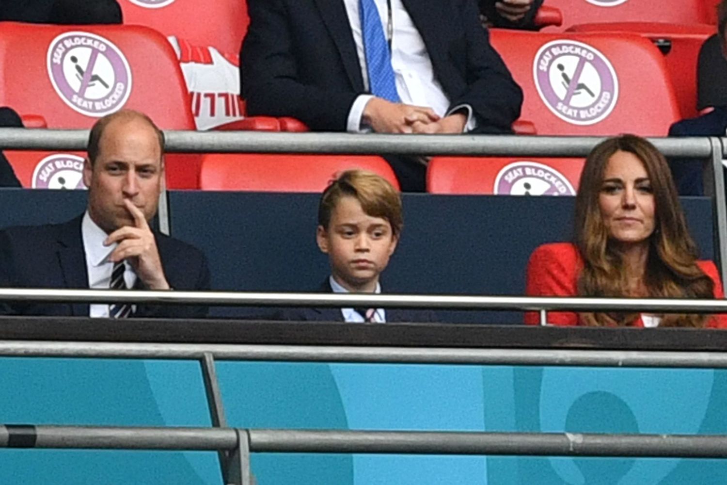 Prince William with his hand on his chin, wearing a suit, sits next to Prince George and Kate Middleton as they watch a soccer game.