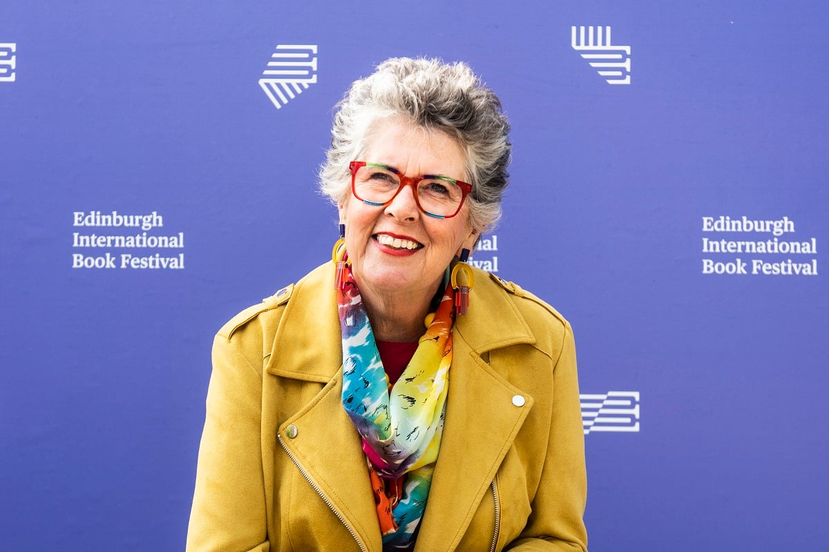 Prue Leith returns to The Great British Baking Show Season 9 -- Prue is wearing a yellow jacket and a rainbow scarf and glasses frames. 