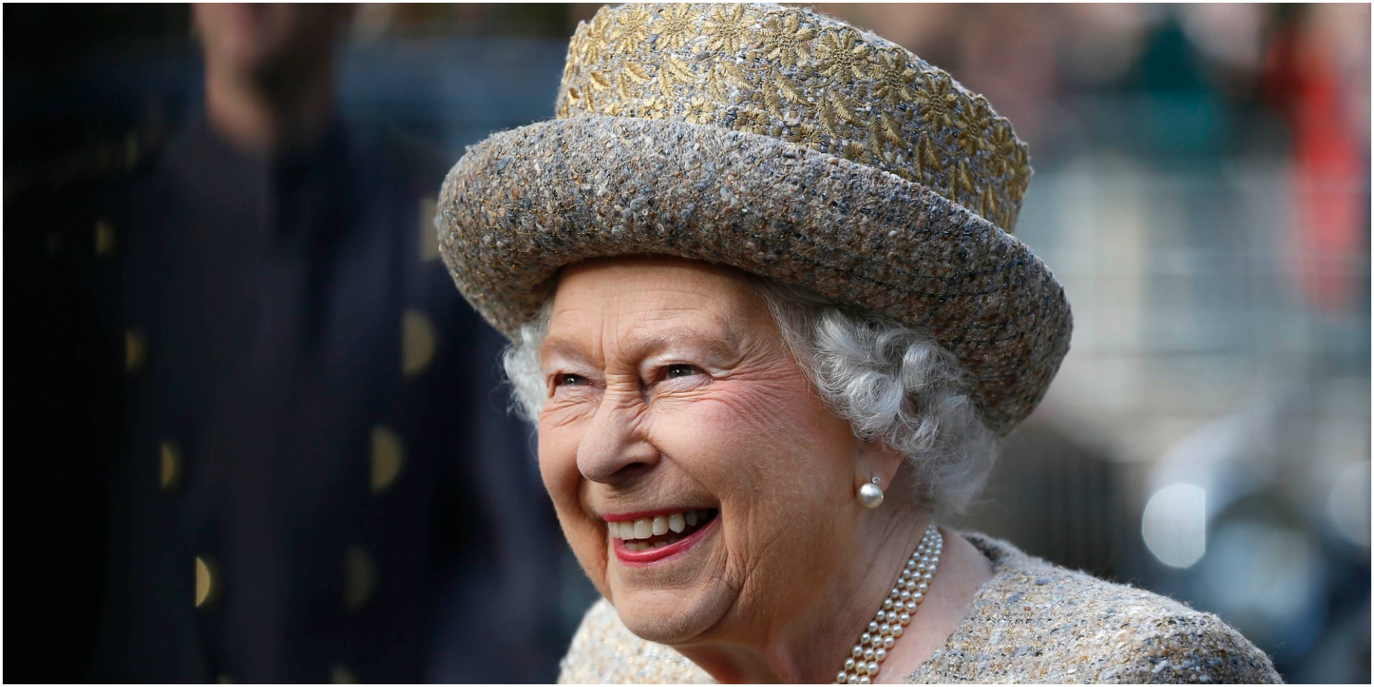 Queen Elizabeth smiles wearing a hat, pink outfit, and pearl necklace