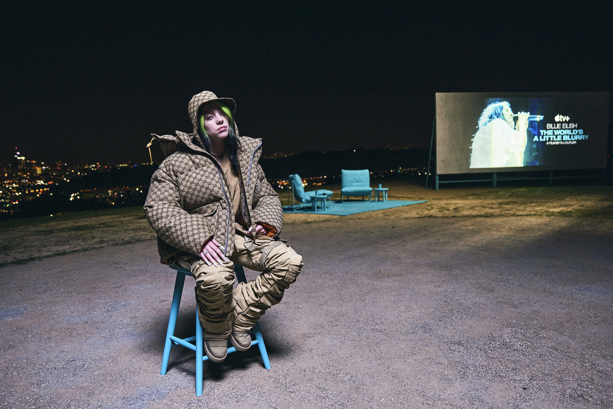 Billie Eilish sits in a blue chair on a hill above Los Angeles for the Apple TV premiere of 'The World's a Little Blurry.'