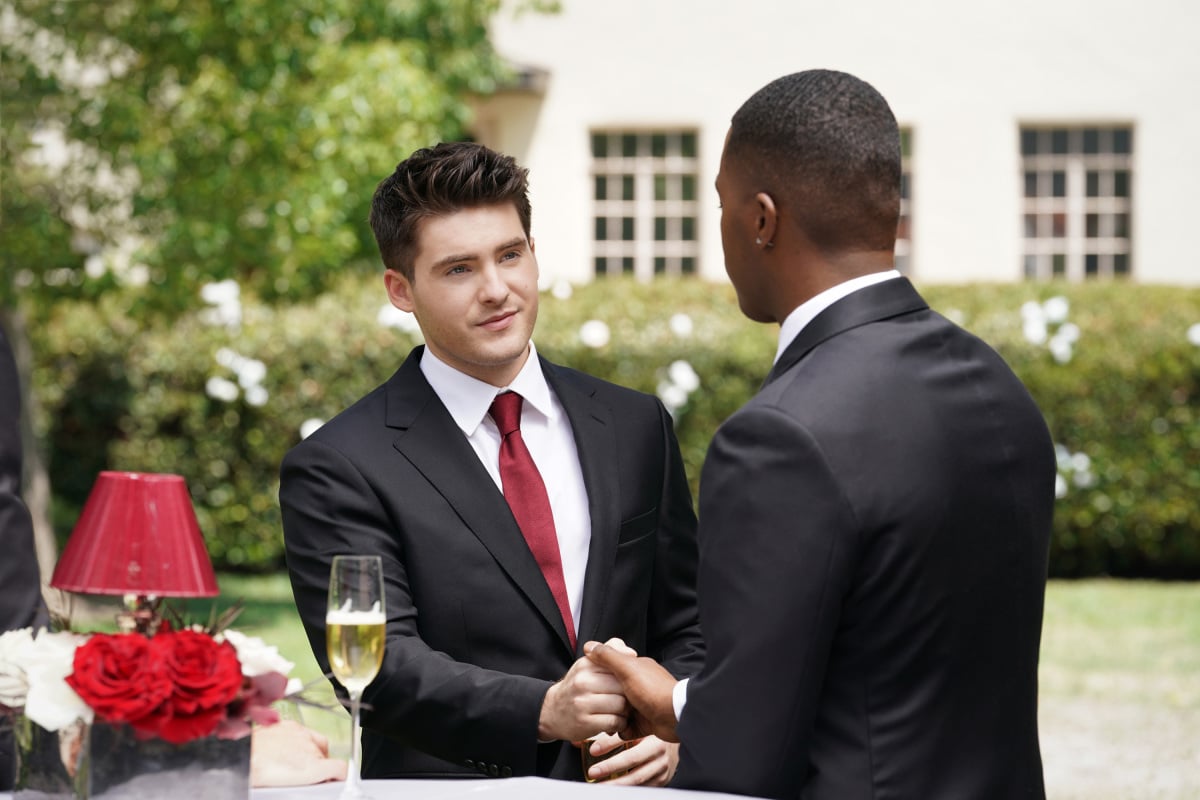 'All American' Season 4 actors Cody Christian and Daniel Ezra, as their characters, shake hands. Both of them are wearing black suits over white shirts and a red tie.