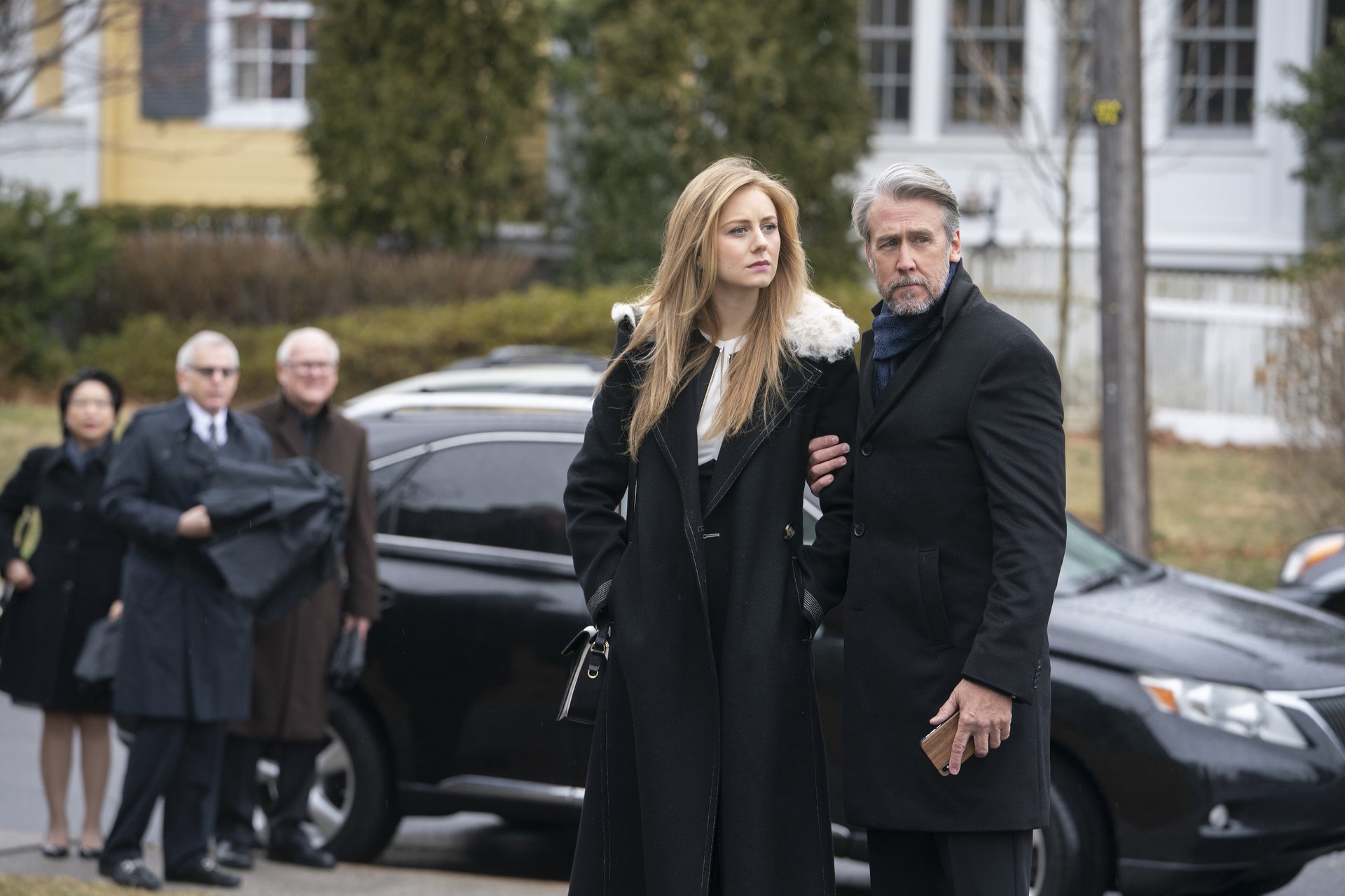 Alan Ruck and Justine Lupe wear black coats and stand outside in Succession.