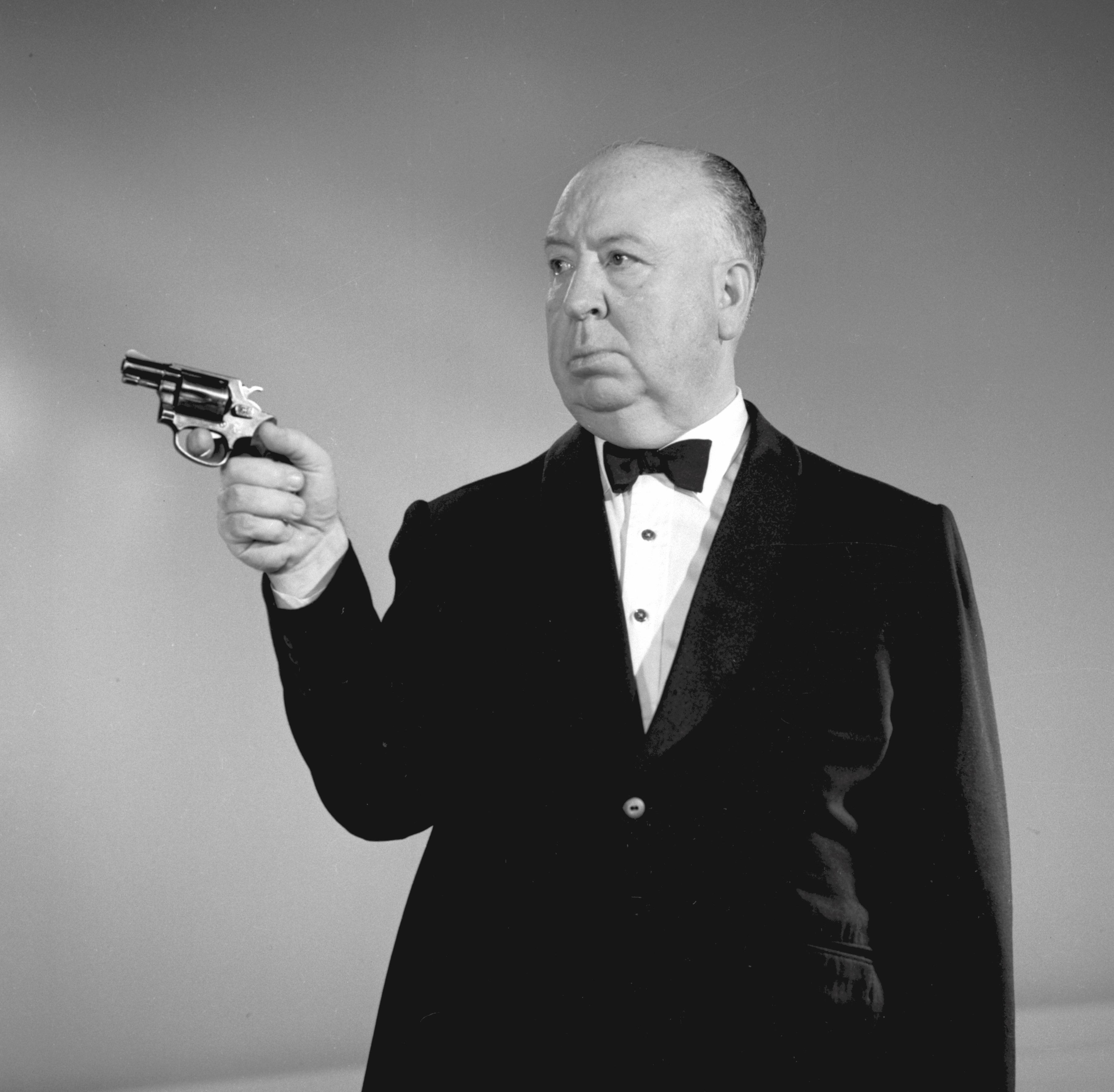 Alfred Hitchcock in a tuxedo holding a gun.