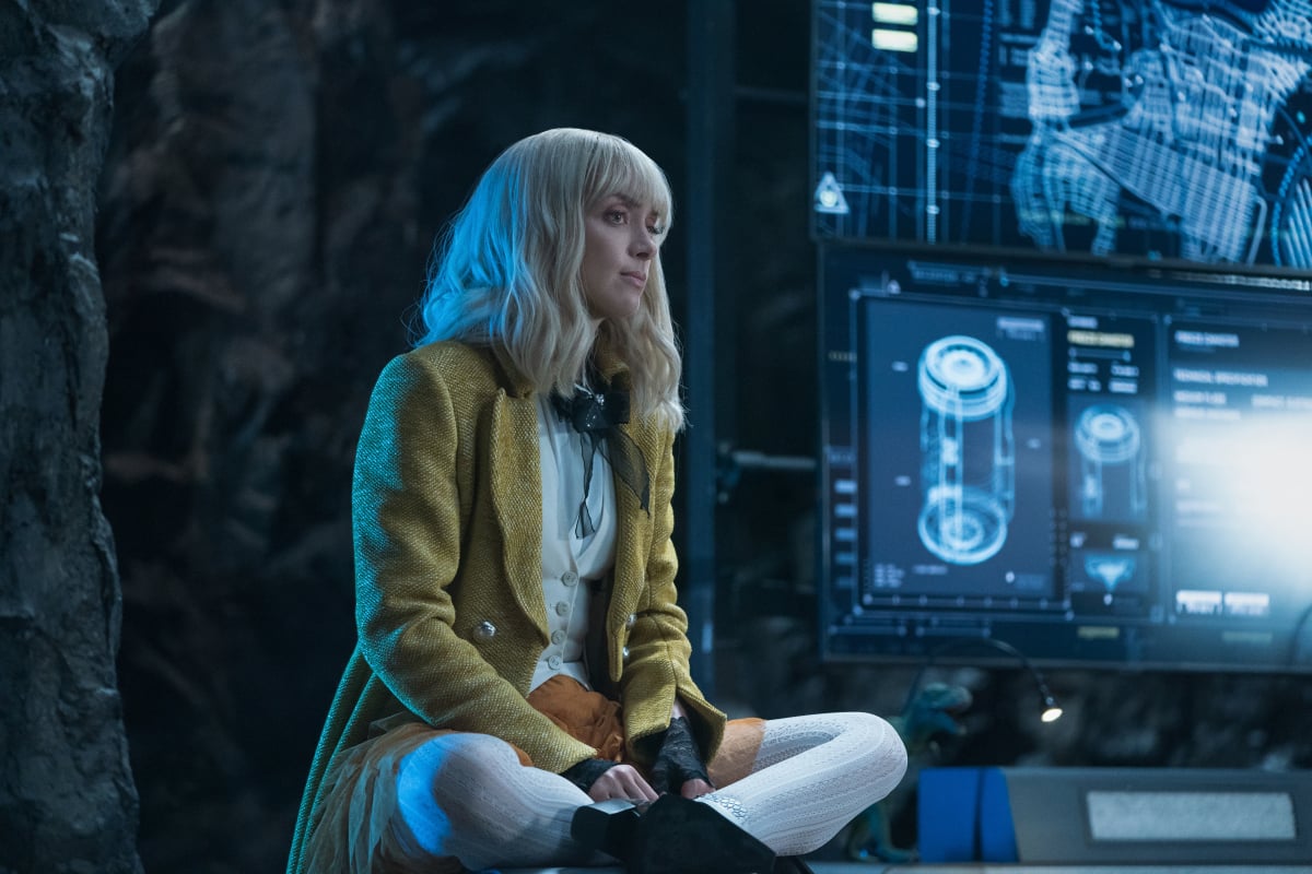 'Batwoman' Season 3 actor Rachel Skarsten, in character as Alice, sits with her legs crossed in the Batcave. She wears a yellow coat, a white vest, brown skirt, and white tights.