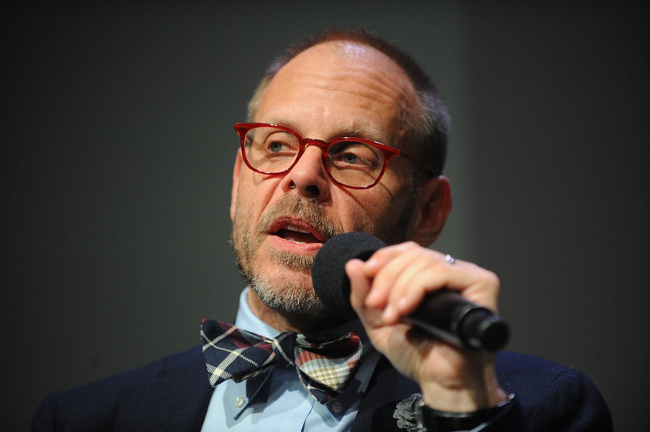 Alton Brown wears glasses and a bow tie as he speaks into a microphone