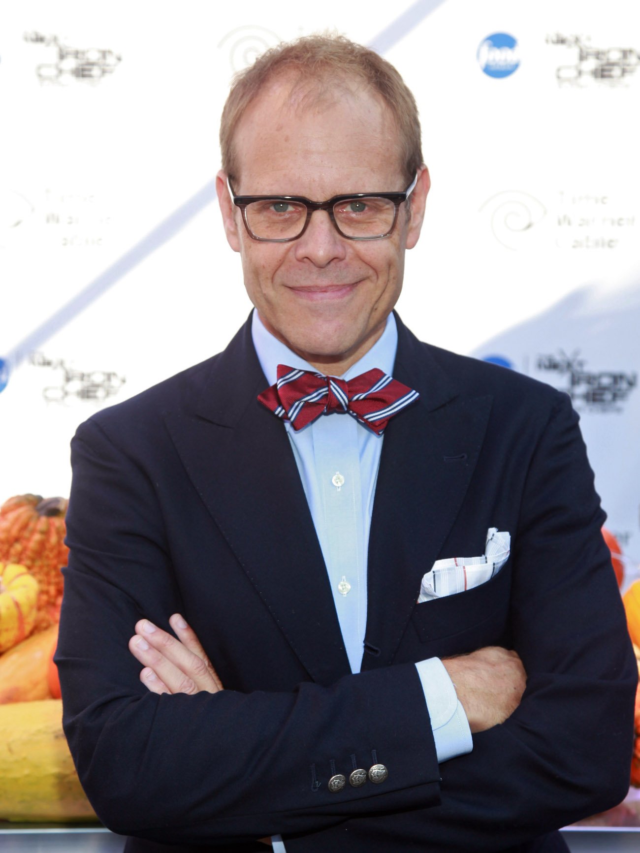 Alton Brown smirks as he stands with his arms folded wearing a blazer and bow tie
