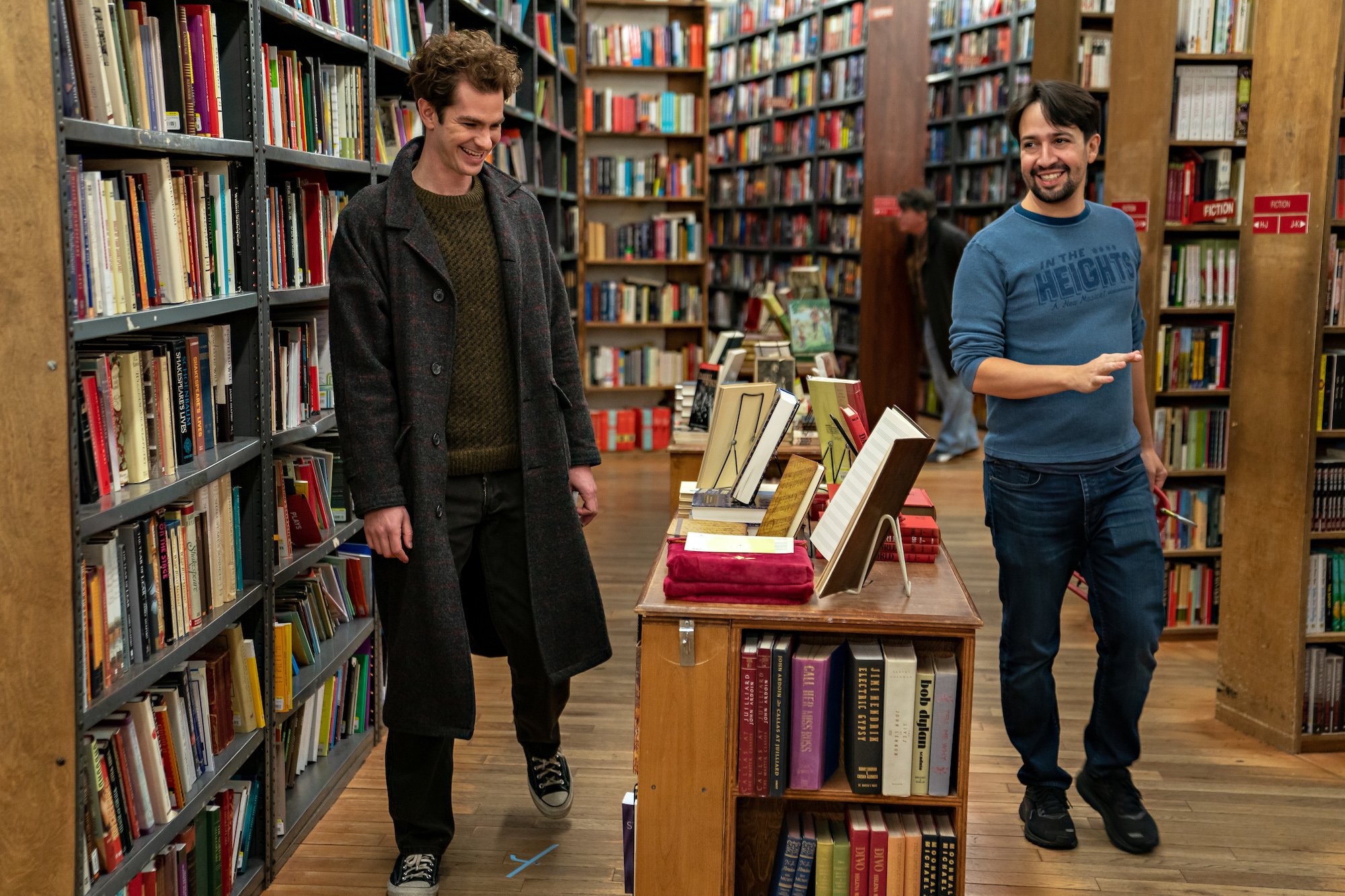 Andrew Garfield and Lin-Manuel Miranda on set of 'Tick, Tick... BOOM!' They walk through a book store smiling. Garfield wears a green sweater, grey wool coat, jeans, and black Converses. Miranda wears a blue long-sleeve shirt that says 'In The Heights' with jeans and sneakers. They're surrounded by large bookshelves filled with books.